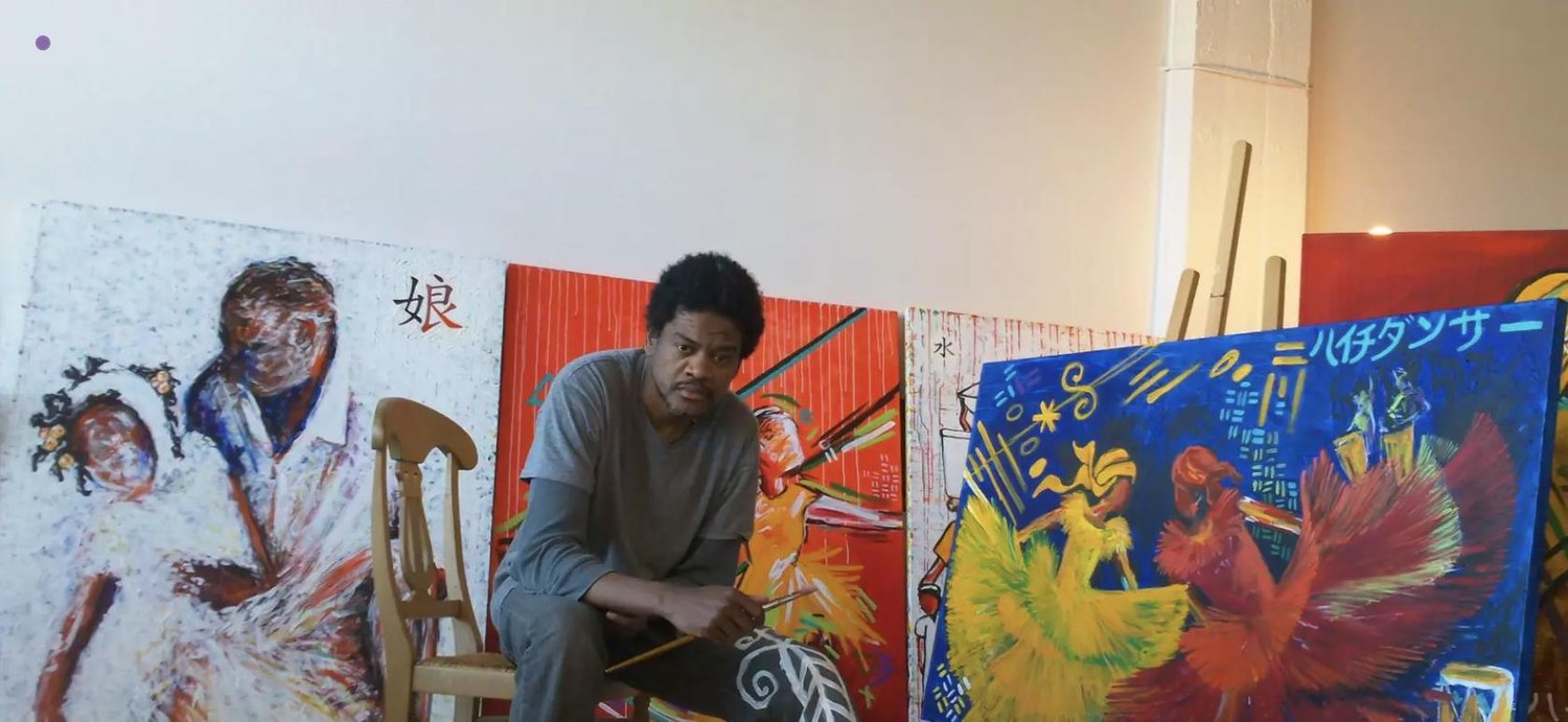 Hertz Nazaire seated in his studio, paintbrush in hand, surrounded by his large canvases